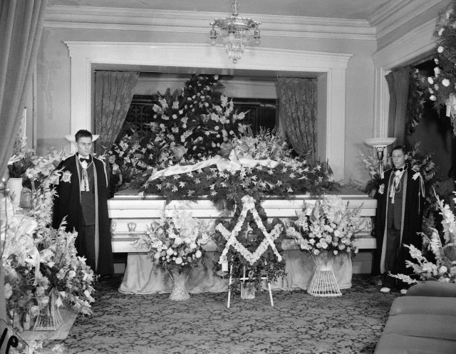 Velório do Robert com dois DeMolays ao lado do caixão e flores em formato de esquadro e compasso.